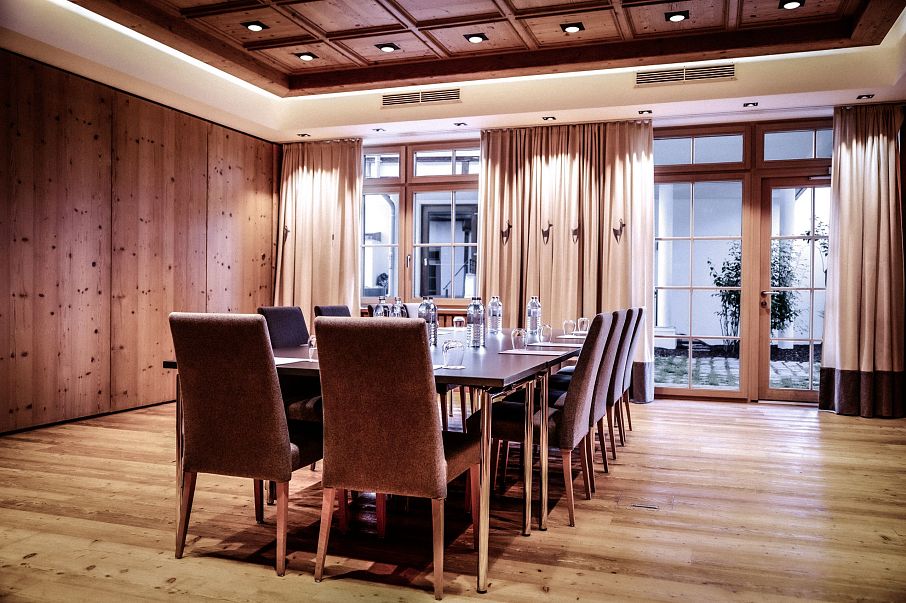 Eleganter, moderner Konferenzraum mit Holzwänden und -boden. Ein rechteckiger Tisch mit acht Stühlen steht im Zentrum. Große Fenster mit Vorhängen im Hintergrund.