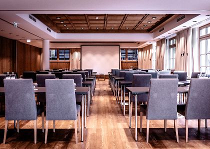 Ein leerer Konferenzraum mit mehreren Reihen von bequemen Stühlen und Tischen, die auf eine große Projektionsleinwand ausgerichtet sind. Holzboden und warme Beleuchtung.