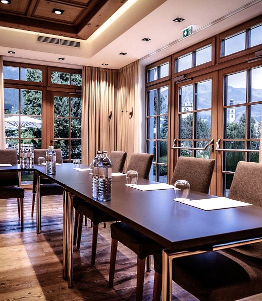 Eleganter Konferenzraum mit Holzboden, großen Fenstern, die viel Tageslicht hereinlassen, und dunklen Holztischen, die mit Wasserkarafeln und Schreibutensilien gedeckt sind.
