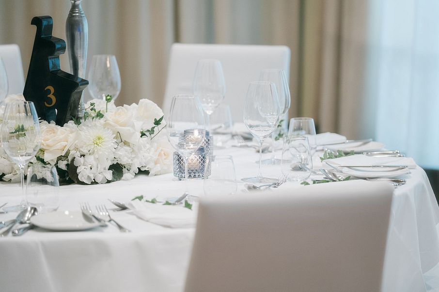 Eleganter, gedeckter Tisch mit weißem Tischtuch und Stühlen. Blumenarrangement und Kerze in der Mitte, umgeben von Weingläsern und Geschirr.