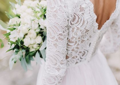 Nahaufnahme einer Frau in einem eleganten, weißen Spitzenhochzeitskleid mit einem tiefen Rückenausschnitt. Sie hält einen Strauß weißer Blumen in der Hand.