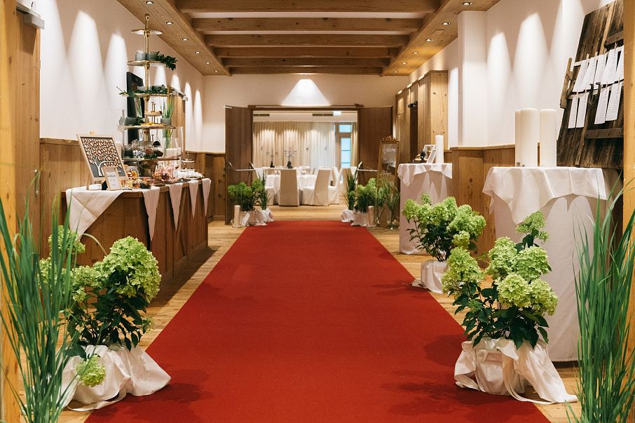 Ein eleganter Veranstaltungsraum mit rotem Teppich, dekoriert mit grünen Pflanzen und weißen Tüchern. Tische säumen die Wände, und sanftes Licht beleuchtet den Raum.