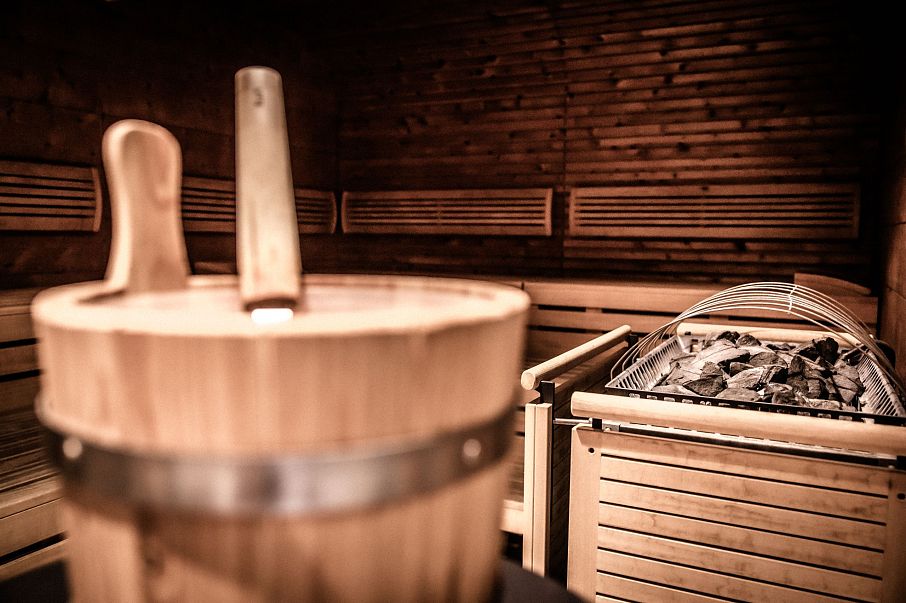 Ein Innenraum einer Sauna mit Holzwänden und Holzbänken. Im Vordergrund ist ein hölzerner Wassereimer mit Schöpflöffel zu sehen. Im Hintergrund liegt die Ofenstelle mit Steinen.