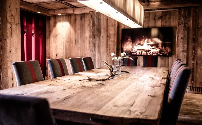 Gemütlicher Speisesaal in rustikalem Stil mit Holzwänden und -decken. Ein langer Holztisch ist mit Stühlen umgeben, im Hintergrund knistert ein Kaminfeuer.