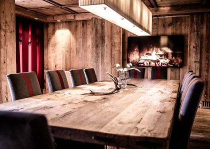 Gemütlicher Speisesaal in rustikalem Stil mit Holzwänden und -decken. Ein langer Holztisch ist mit Stühlen umgeben, im Hintergrund knistert ein Kaminfeuer.