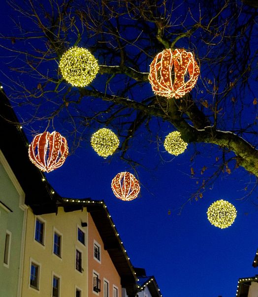 Ein abendlicher Blick auf eine festlich beleuchtete Straße mit farbenfrohen Häuserfassaden und leuchtenden Kugeln, die an den Ästen eines Baumes hängen.