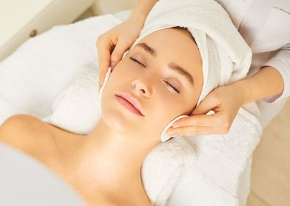 Junge Frau genießt eine Gesichtsbehandlung in einem Spa. Ihr Kopf ist in ein Handtuch gewickelt und ihre Haut wird sanft mit Wattepads behandelt.