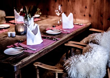 Gemütlicher rustikaler Esstisch mit flauschigen Fellstühlen, lila Platzsets, weiß gefalteten Servietten und elegantem Geschirr. Dezente Beleuchtung.