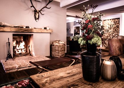Gemütliches Wohnzimmer mit Kaminfeuer, rustikaler Wood-Tisch, Vase mit Blumenarrangement und Geweihdekor an der Wand. Einladende und warme Atmosphäre.