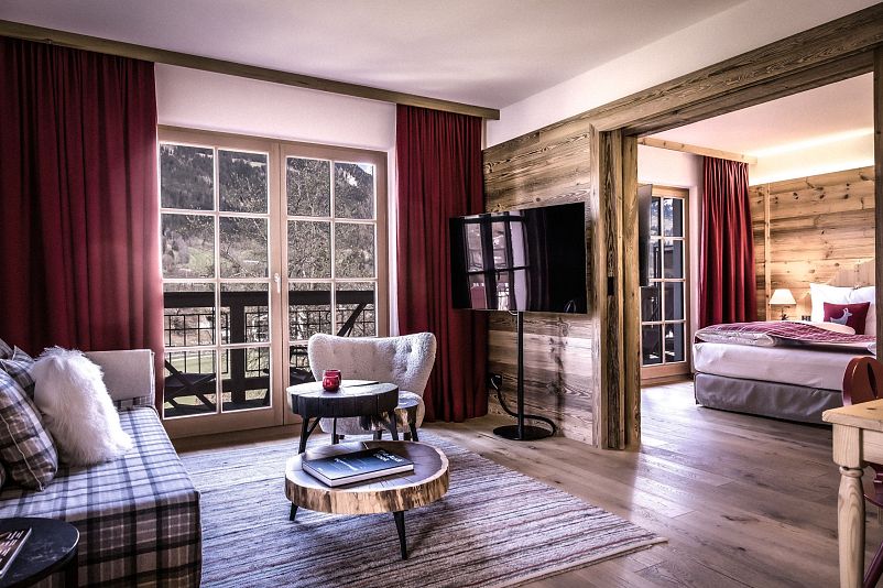 Gemütliche Hotelsuite im alpinen Stil mit Holzmöbeln, roten Vorhängen und modernen Annehmlichkeiten. Schlafzimmer und Wohnzimmer durch Holztrennwand getrennt.