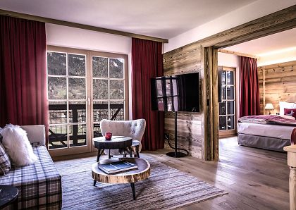 Gemütliche Hotelsuite im alpinen Stil mit Holzmöbeln, roten Vorhängen und modernen Annehmlichkeiten. Schlafzimmer und Wohnzimmer durch Holztrennwand getrennt.