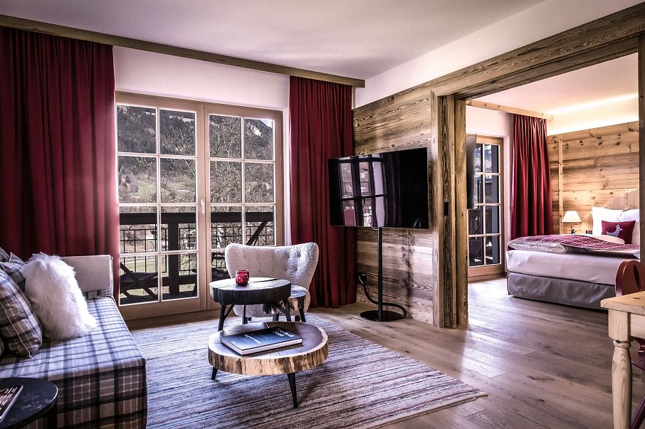 Gemütliche Hotelsuite im alpinen Stil mit Holzmöbeln, roten Vorhängen und modernen Annehmlichkeiten. Schlafzimmer und Wohnzimmer durch Holztrennwand getrennt.