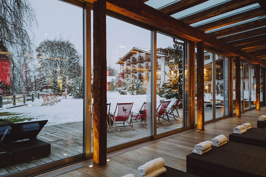 Ein gemütlicher Wellnessbereich mit großen Glasfenstern bietet Aussicht auf eine verschneite Landschaft mit gemütlichen Liegestühlen und einem Holzhotel im Hintergrund.