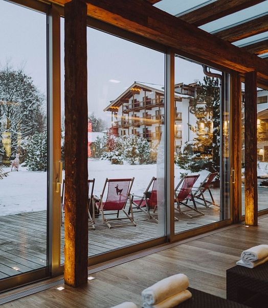 Ein gemütlicher Wellnessbereich mit großen Glasfenstern bietet Aussicht auf eine verschneite Landschaft mit gemütlichen Liegestühlen und einem Holzhotel im Hintergrund.