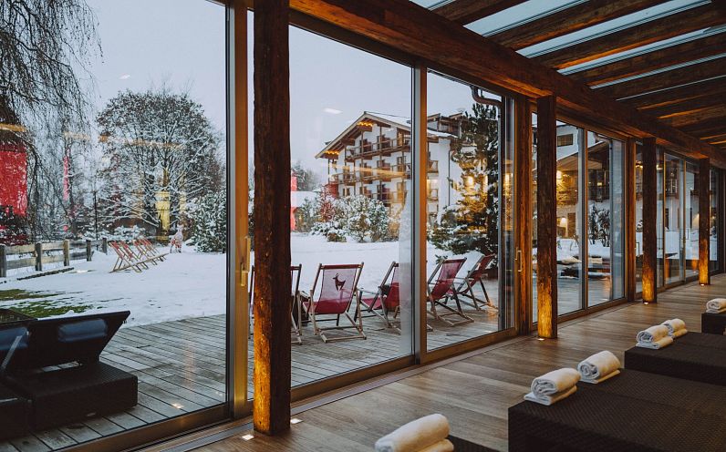 Ein gemütlicher Wellnessbereich mit großen Glasfenstern bietet Aussicht auf eine verschneite Landschaft mit gemütlichen Liegestühlen und einem Holzhotel im Hintergrund.