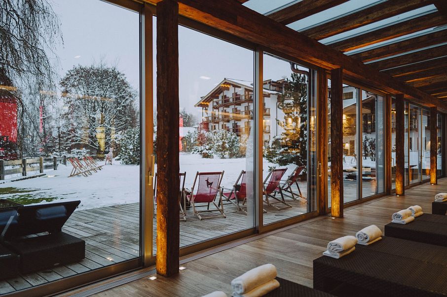 Ein gemütlicher Wellnessbereich mit großen Glasfenstern bietet Aussicht auf eine verschneite Landschaft mit gemütlichen Liegestühlen und einem Holzhotel im Hintergrund.