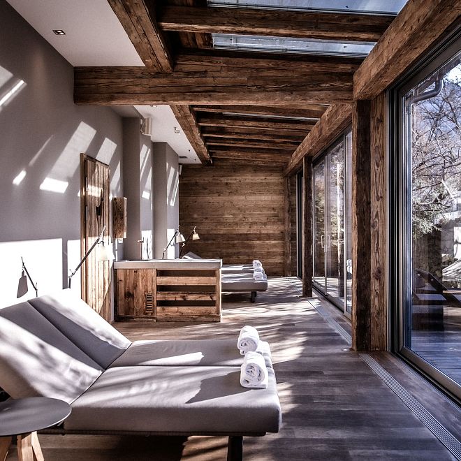 Gemütlicher Wellnessraum mit Holzdetails, Liegestühlen und großen Fenstern, die natürliche Lichtstrahlen hineinlassen. Berglandschaft ist draußen sichtbar.