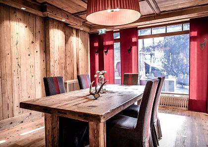 Gemütlicher Essbereich im Alpenstil mit Holzwänden und -decke, rustikalem Holztisch, sechs lederbezogenen Stühlen und rotem Vorhang an einem großen Fenster.