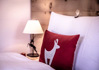 Ein gemütliches Schlafzimmer mit einem weißen Bettbezug und einem roten Kissen mit Hirschmotiv. Eine Tischlampe mit Tierfuß steht auf einem Holztisch daneben.