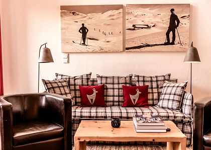 Gemütliches Wohnzimmer mit kariertem Sofa, roten Kissen, zwei Ledersesseln und einem Holztisch. An der Wand hängen Bilder von Skifahrern im Schnee.