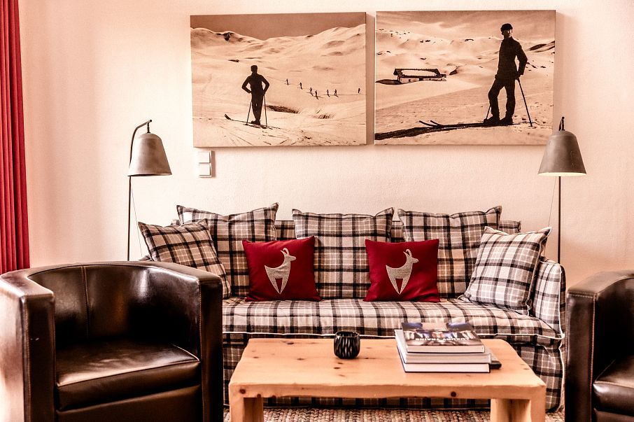 Gemütliches Wohnzimmer mit kariertem Sofa, roten Kissen, zwei Ledersesseln und einem Holztisch. An der Wand hängen Bilder von Skifahrern im Schnee.
