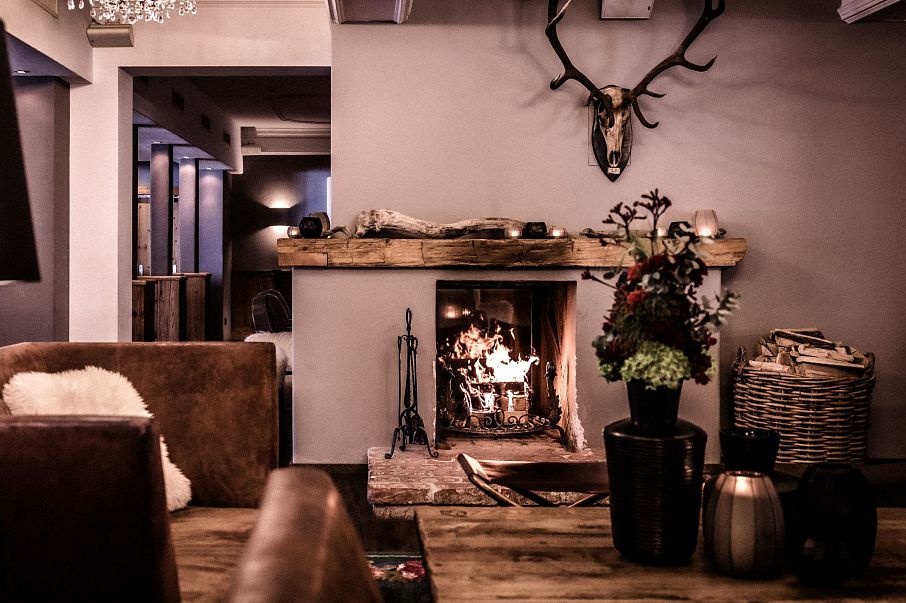 Gemütlicher Wohnraum mit Kamin, bequemen Sesseln und Sofas. Warme Beleuchtung, Holzverkleidung und Kunstwerk an der Wand schaffen eine einladende Atmosphäre.