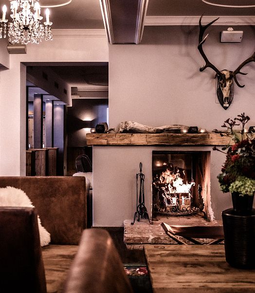 Gemütlicher Wohnraum mit Kamin, bequemen Sesseln und Sofas. Warme Beleuchtung, Holzverkleidung und Kunstwerk an der Wand schaffen eine einladende Atmosphäre.