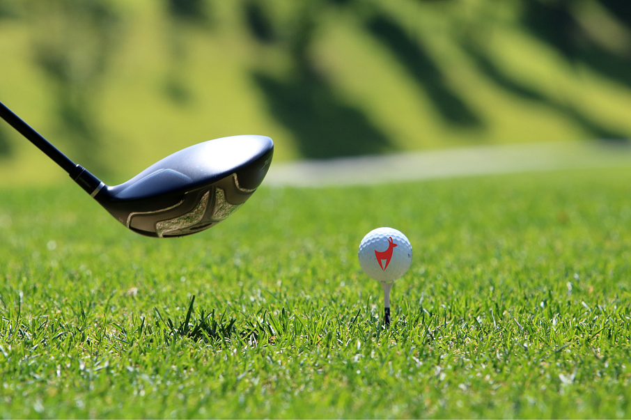 Nahaufnahme eines Golfschlägers, der bereit ist, einen weißen Golfball auf einem Tee zu schlagen. Der grüne Rasen und unscharfe Hintergründe sind sichtbar.