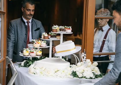 Zwei Männer in Anzügen bringen eine mehrstöckige Torte und Cupcakes auf einem dekorierten Tisch in einen Raum. Weiße Blumen schmücken den Tisch.