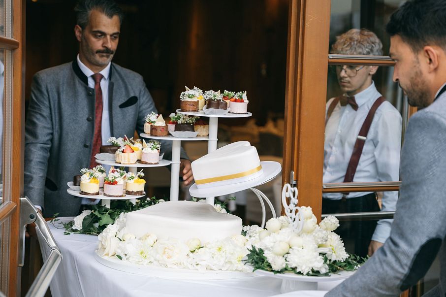 Zwei Männer in Anzügen bringen eine mehrstöckige Torte und Cupcakes auf einem dekorierten Tisch in einen Raum. Weiße Blumen schmücken den Tisch.