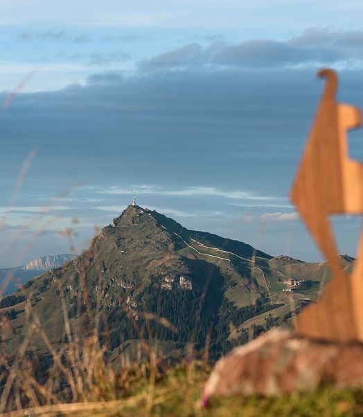 Holzskulptur steht im Vordergrund vor einer malerischen Berglandschaft mit sanften Hügeln und bewölktem Himmel. Der Gipfel eines Berges ist im Hintergrund zu sehen.