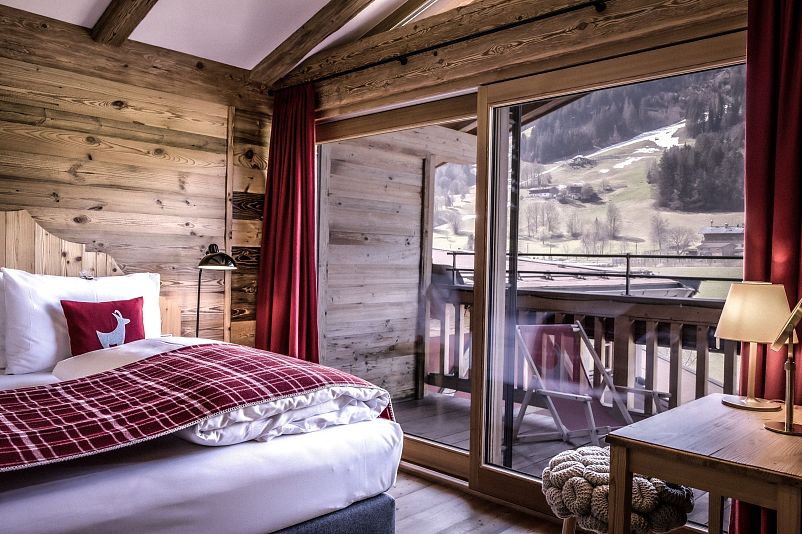 Gemütliches Schlafzimmer aus Holz mit großem Fenster und Aussicht auf verschneite Berge. Rotes Dekor, modernes Bett und ein Balkon runden den Raum ab.