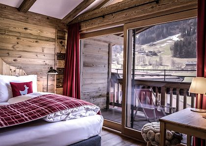 Gemütliches Schlafzimmer aus Holz mit großem Fenster und Aussicht auf verschneite Berge. Rotes Dekor, modernes Bett und ein Balkon runden den Raum ab.