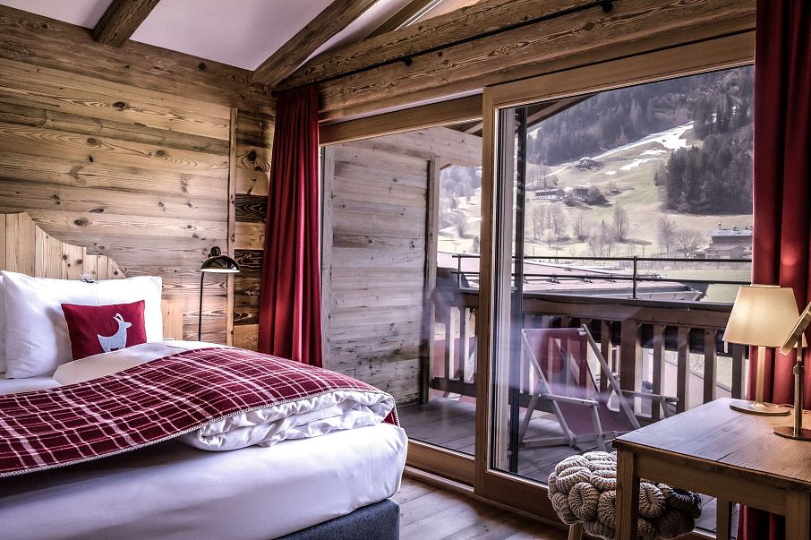 Gemütliches Schlafzimmer aus Holz mit großem Fenster und Aussicht auf verschneite Berge. Rotes Dekor, modernes Bett und ein Balkon runden den Raum ab.