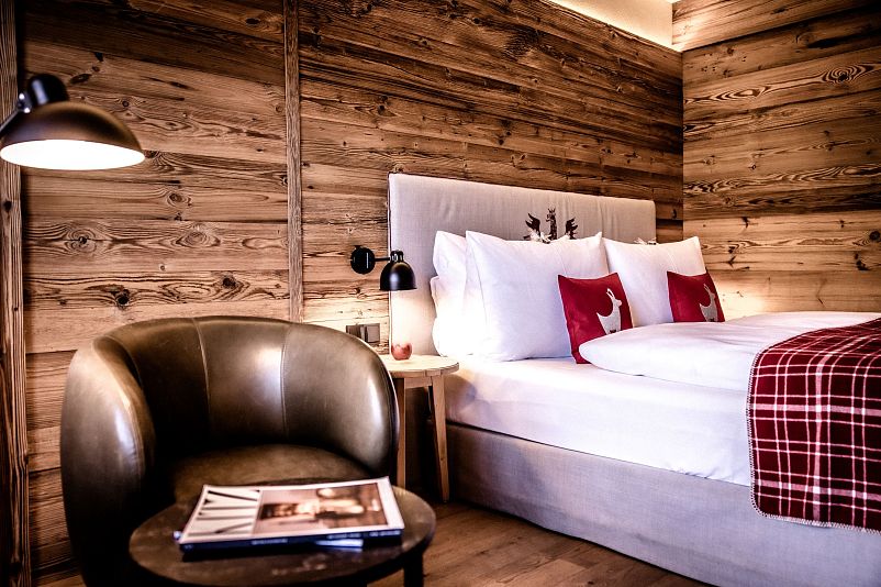 Gemütliches Schlafzimmer mit Holzwänden, einem großen Bett mit weißen Kissen und roten Hirschkissen. Ein Ledersessel steht neben einem runden Tisch mit Zeitschriften.