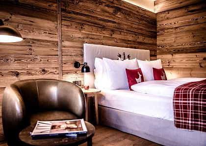 Gemütliches Schlafzimmer mit Holzwänden, einem großen Bett mit weißen Kissen und roten Hirschkissen. Ein Ledersessel steht neben einem runden Tisch mit Zeitschriften.