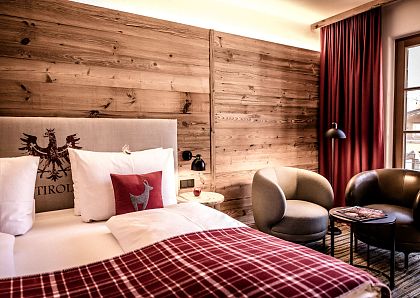 Gemütliches Hotelzimmer mit Holzwänden, rotem Vorhang und Tagesdecke mit kariertem Muster. Zwei graue Sessel und ein kleiner Tisch vervollständigen das Ambiente.