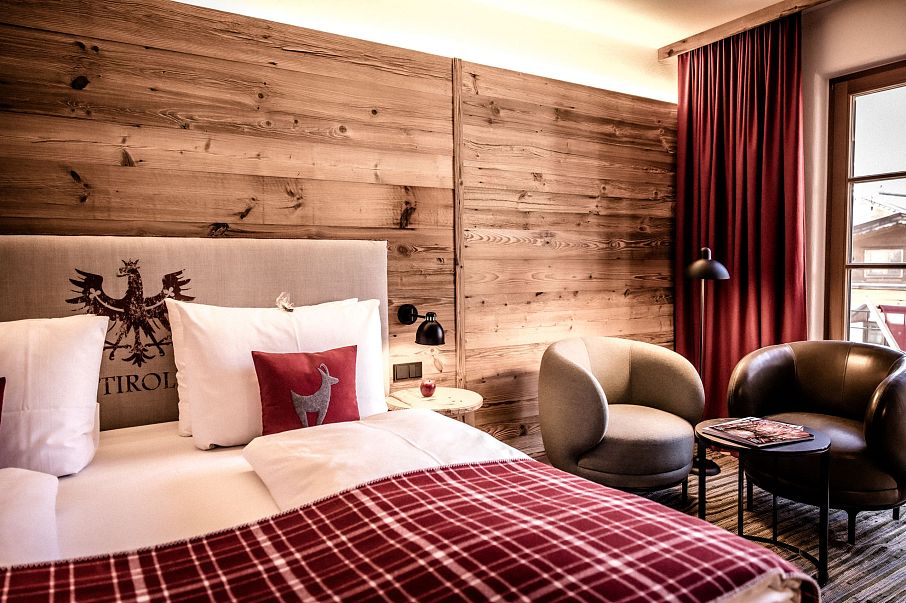 Gemütliches Hotelzimmer mit Holzwänden, rotem Vorhang und Tagesdecke mit kariertem Muster. Zwei graue Sessel und ein kleiner Tisch vervollständigen das Ambiente.