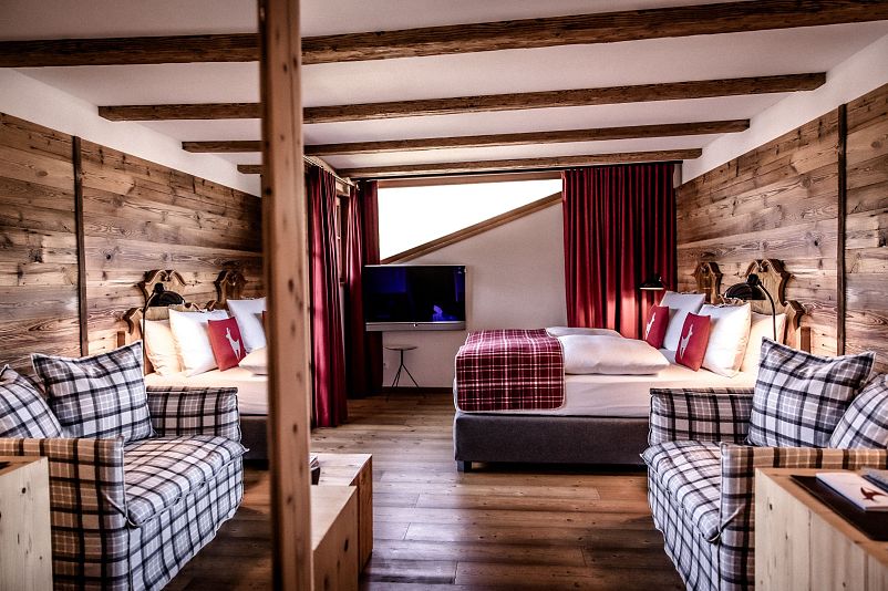 Gemütliches Chalet-Zimmer mit Holzwänden und freiliegenden Deckenbalken, ausgestattet mit einem Doppelbett, zwei Sofas, einem Fernseher und roten Vorhängen.