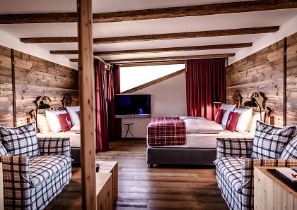 Gemütliches Chalet-Zimmer mit Holzwänden und freiliegenden Deckenbalken, ausgestattet mit einem Doppelbett, zwei Sofas, einem Fernseher und roten Vorhängen.