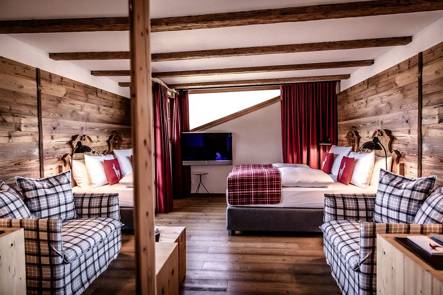 Gemütliches Chalet-Zimmer mit Holzwänden und freiliegenden Deckenbalken, ausgestattet mit einem Doppelbett, zwei Sofas, einem Fernseher und roten Vorhängen.