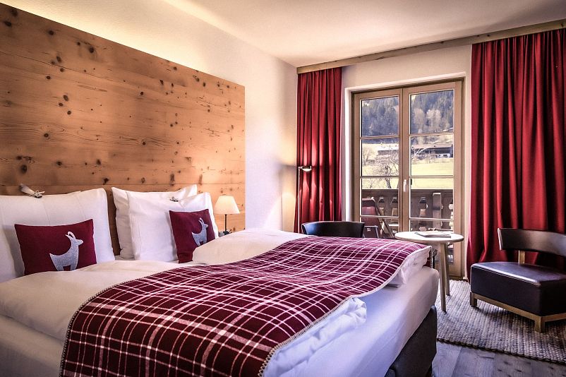Gemütliches Hotelzimmer mit zwei Einzelbetten, roten karierten Decken und Kissen mit Hirschmuster. Holzwandverkleidung, roter Vorhang, Fenster mit Bergblick.