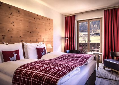 Gemütliches Hotelzimmer mit zwei Einzelbetten, roten karierten Decken und Kissen mit Hirschmuster. Holzwandverkleidung, roter Vorhang, Fenster mit Bergblick.