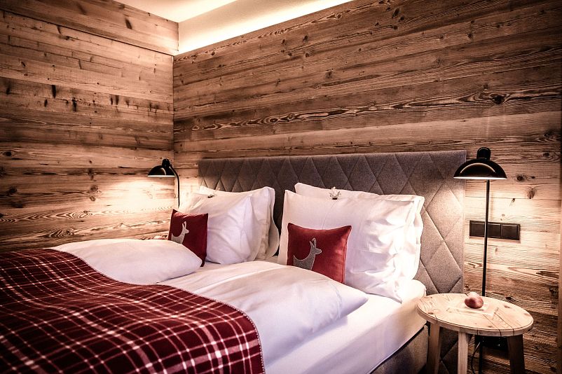 Gemütliches Schlafzimmer mit Holzwänden und warmem Licht. Ein breites Bett ist mit karierten Decken und Kissen mit Hirschmotiven dekoriert. Zwei Lampen beleuchten den Raum.