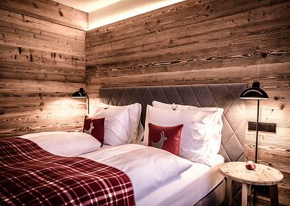 Gemütliches Schlafzimmer mit Holzwänden und warmem Licht. Ein breites Bett ist mit karierten Decken und Kissen mit Hirschmotiven dekoriert. Zwei Lampen beleuchten den Raum.