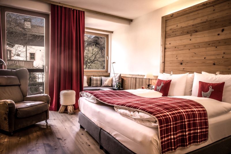 Gemütliches Schlafzimmer mit Holzwänden, einem großen Doppelbett mit roten Karomustern, Plüschsessel und großen Fenstern mit roten Vorhängen.