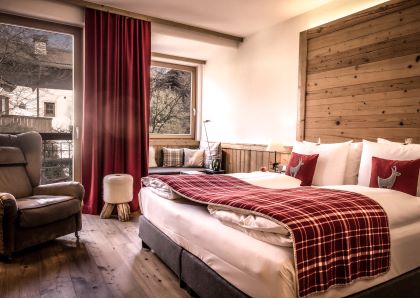 Gemütliches Schlafzimmer mit Holzwänden, einem großen Doppelbett mit roten Karomustern, Plüschsessel und großen Fenstern mit roten Vorhängen.