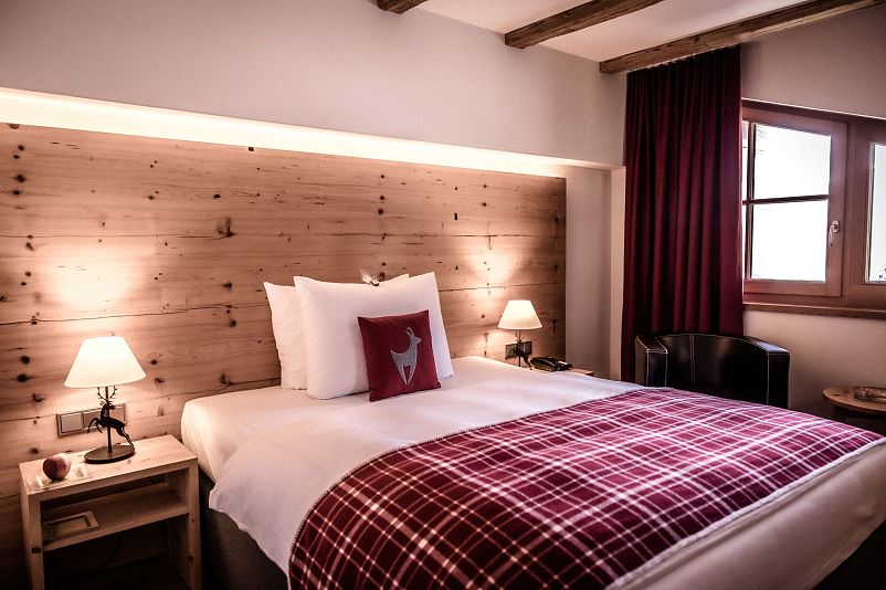Gemütliches Hotelzimmer mit rustikalem Holzwandpaneel, einem großen Doppelbett mit rot-weißer Bettdecke und Kissen, zwei Nachttischlampen und einem Fenster.