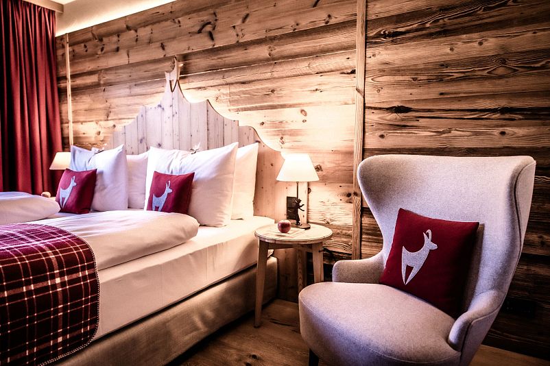 Gemütliches Hotelzimmer mit Holzwänden und rustikalem Charme. Ein großes Bett mit Hirschkissen und ein weicher Sessel schaffen eine warme, einladende Atmosphäre.