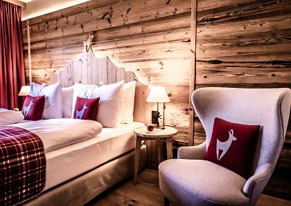 Gemütliches Hotelzimmer mit Holzwänden und rustikalem Charme. Ein großes Bett mit Hirschkissen und ein weicher Sessel schaffen eine warme, einladende Atmosphäre.